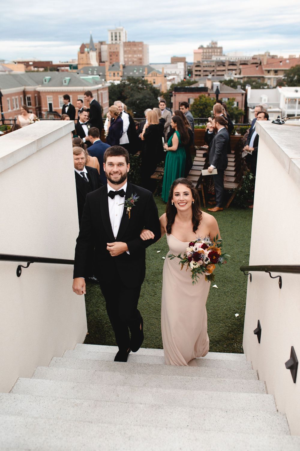 A groomsman and bridesmaid descend rooftop stairs at an outdoor wedding ceremony, she holds a colorful bouquet.