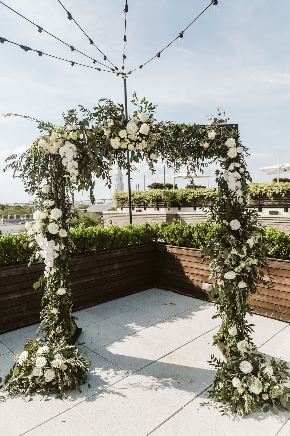 Elegant outdoor wedding arch adorned with white roses and lush greenery on a rooftop venue under string lights.