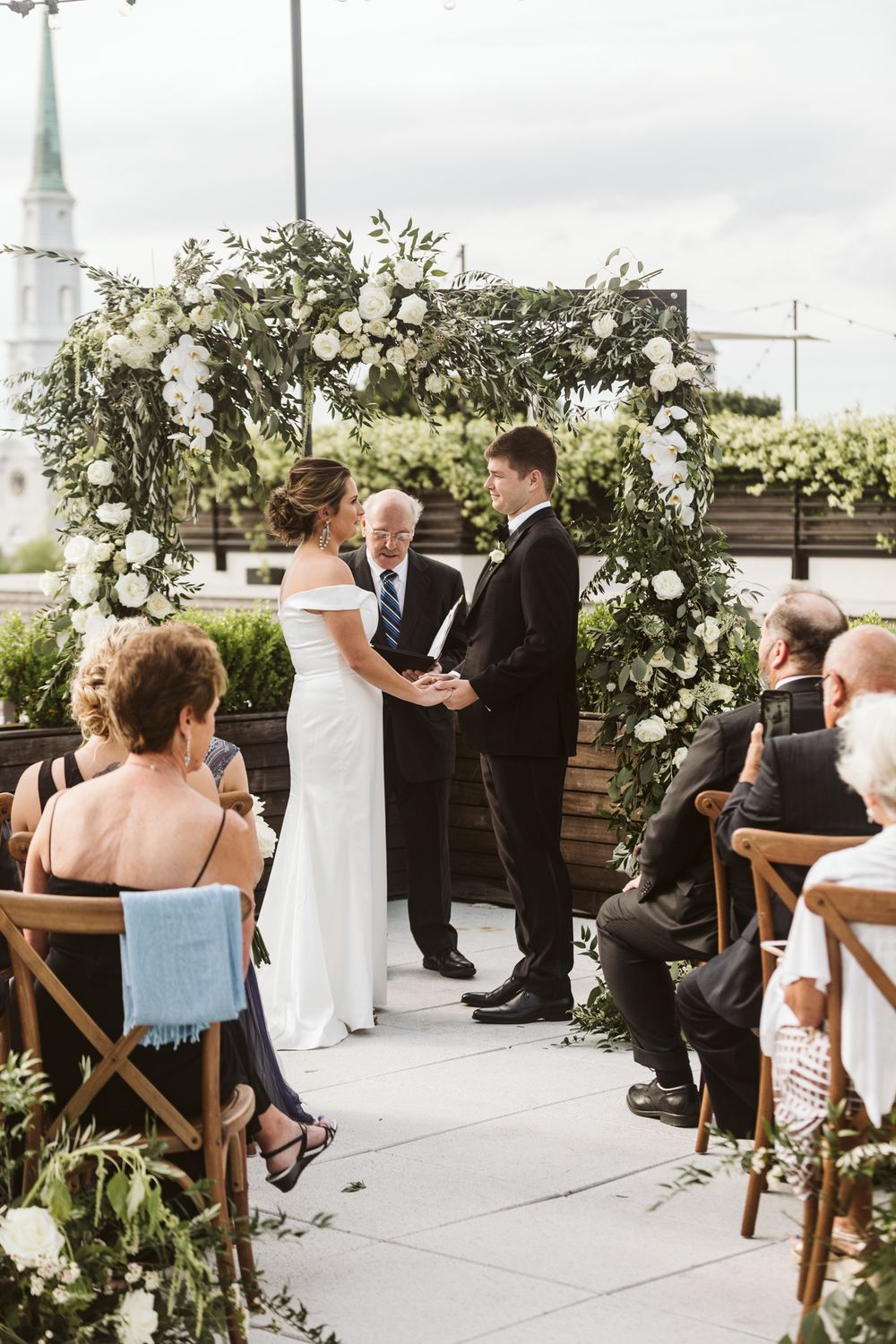 Couple exchanges vows under floral arch with white roses and greenery at outdoor rooftop wedding ceremony.