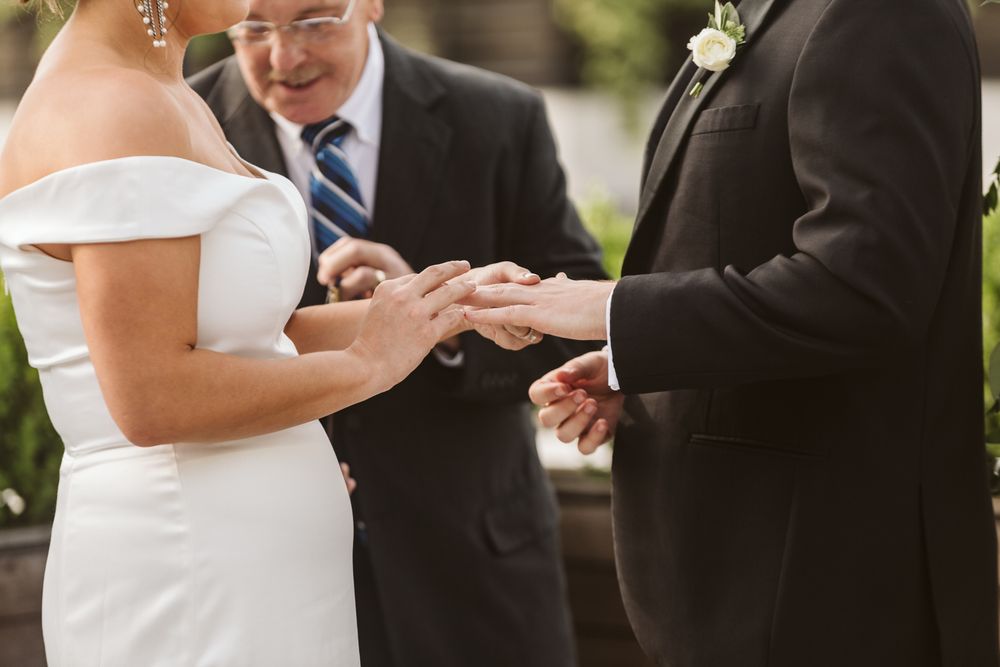 Groom placing wedding ring on bride's finger during ceremony, officiant in background.