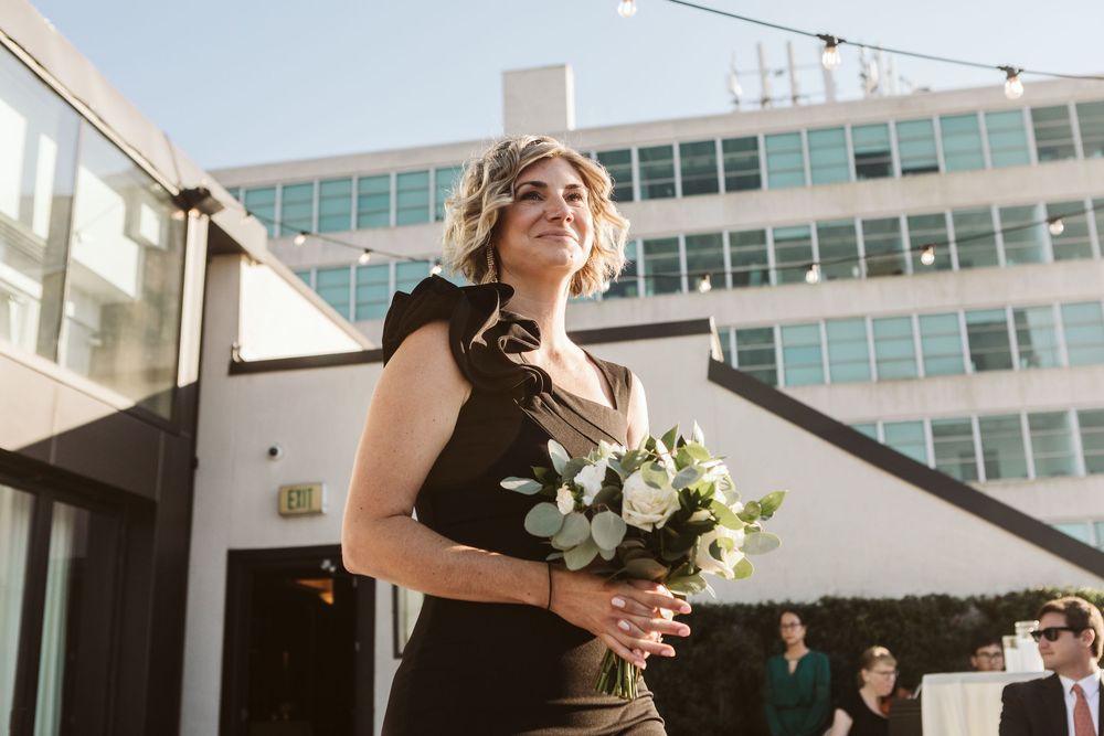 Woman in black dress holding white bouquet outdoors at rooftop wedding venue with modern building backdrop.