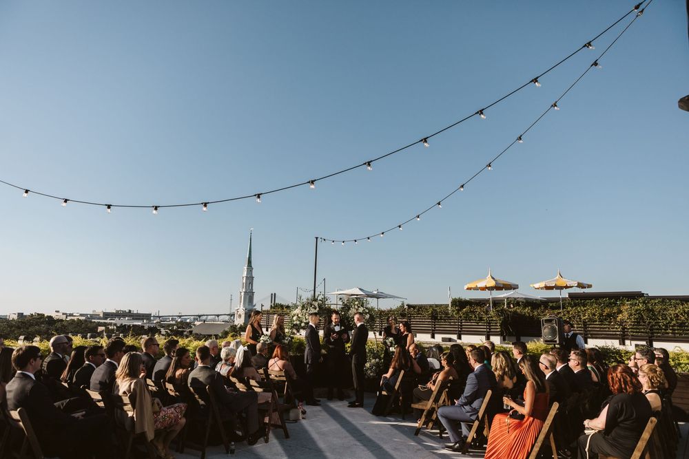 Outdoor rooftop wedding ceremony with string lights, seated guests, and a church spire under clear blue sky.