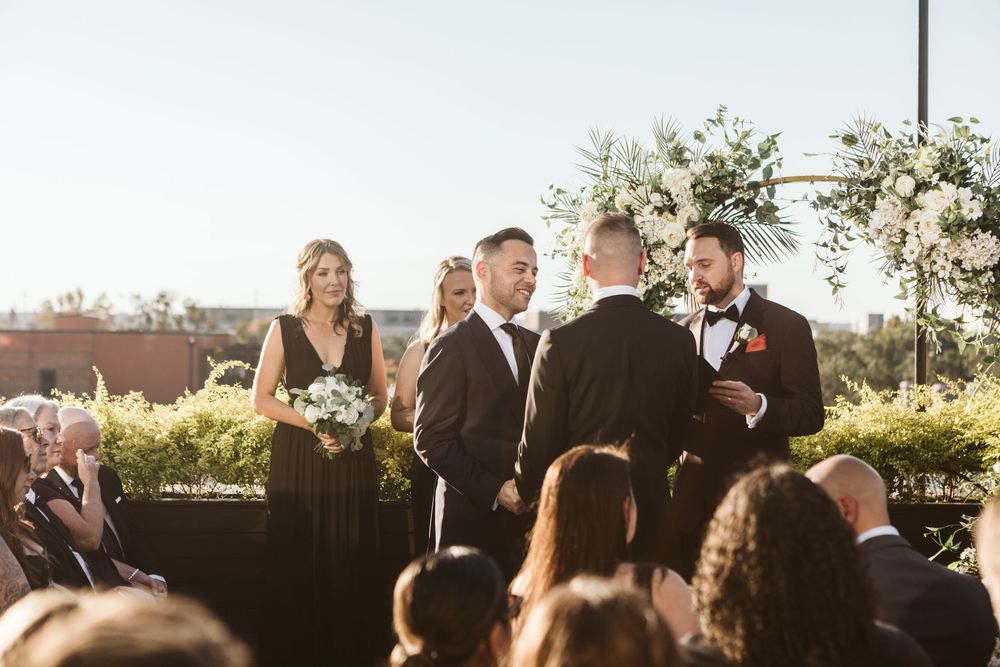 Two grooms exchange vows at an outdoor wedding ceremony with bridesmaids in black dresses and floral arch backdrop.