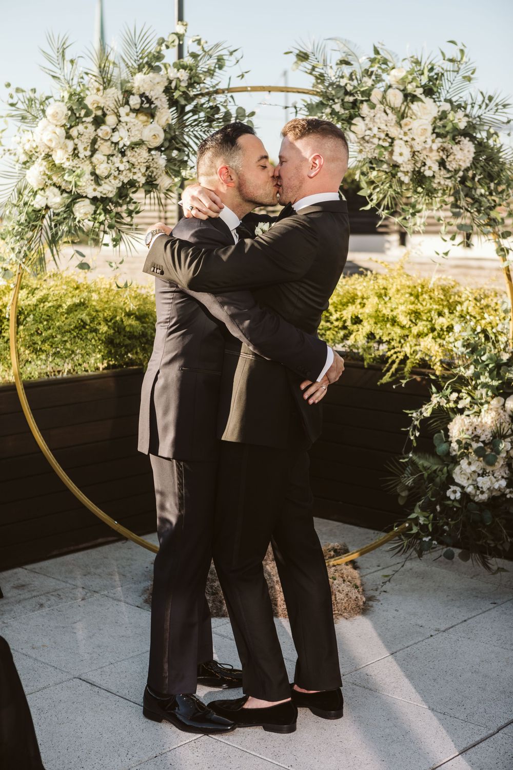 Two grooms share a first kiss at their wedding ceremony in front of a floral arch with white flowers.