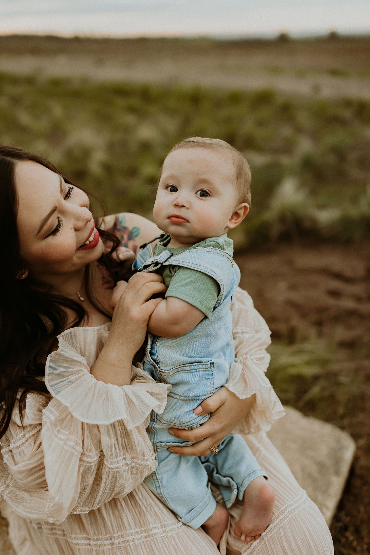 A smiling mother with tattoos holds her baby boy in denim overalls outdoors in a field at golden hour.
Image by Katie Denton