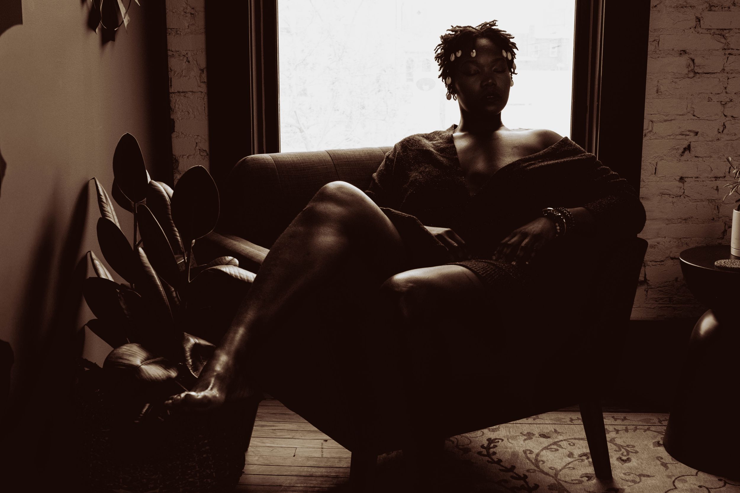 Silhouetted figure sits in an armchair by a bright window in a moody sepia portrait.