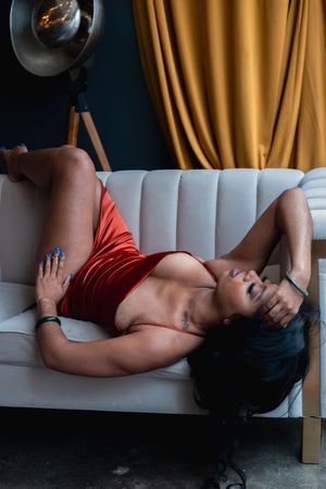 Woman in red bodysuit poses reclined on a white sofa with golden curtains backdrop.