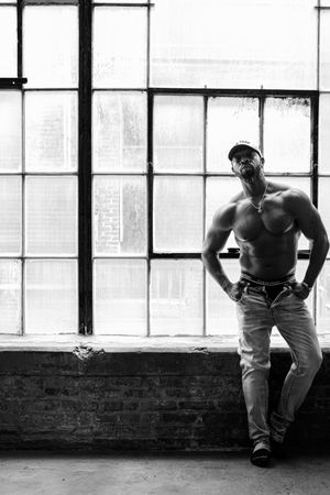 Muscular shirtless man in jeans poses confidently before large industrial windows in black and white.