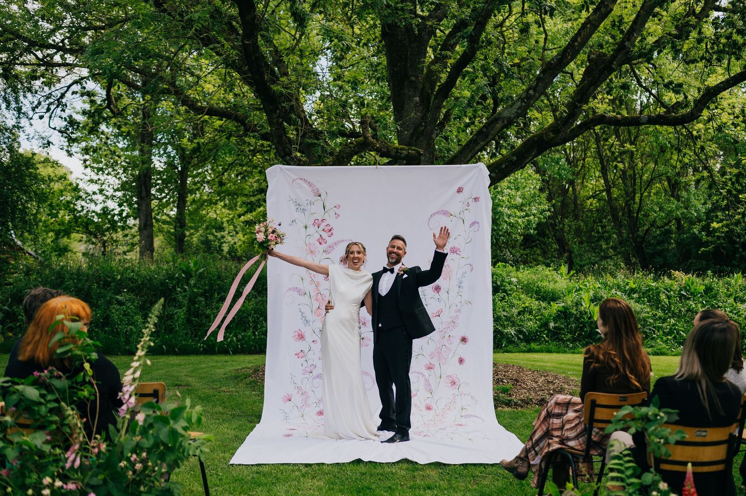 Bride and groom celebrate outdoors in front of a floral white backdrop surrounded by lush green trees at a garden wedding.