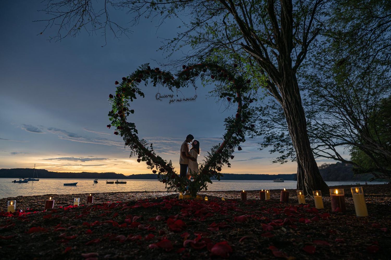 Couple kissing inside a heart-shaped floral wreath at sunset by a lake, surrounded by candles and rose petals.