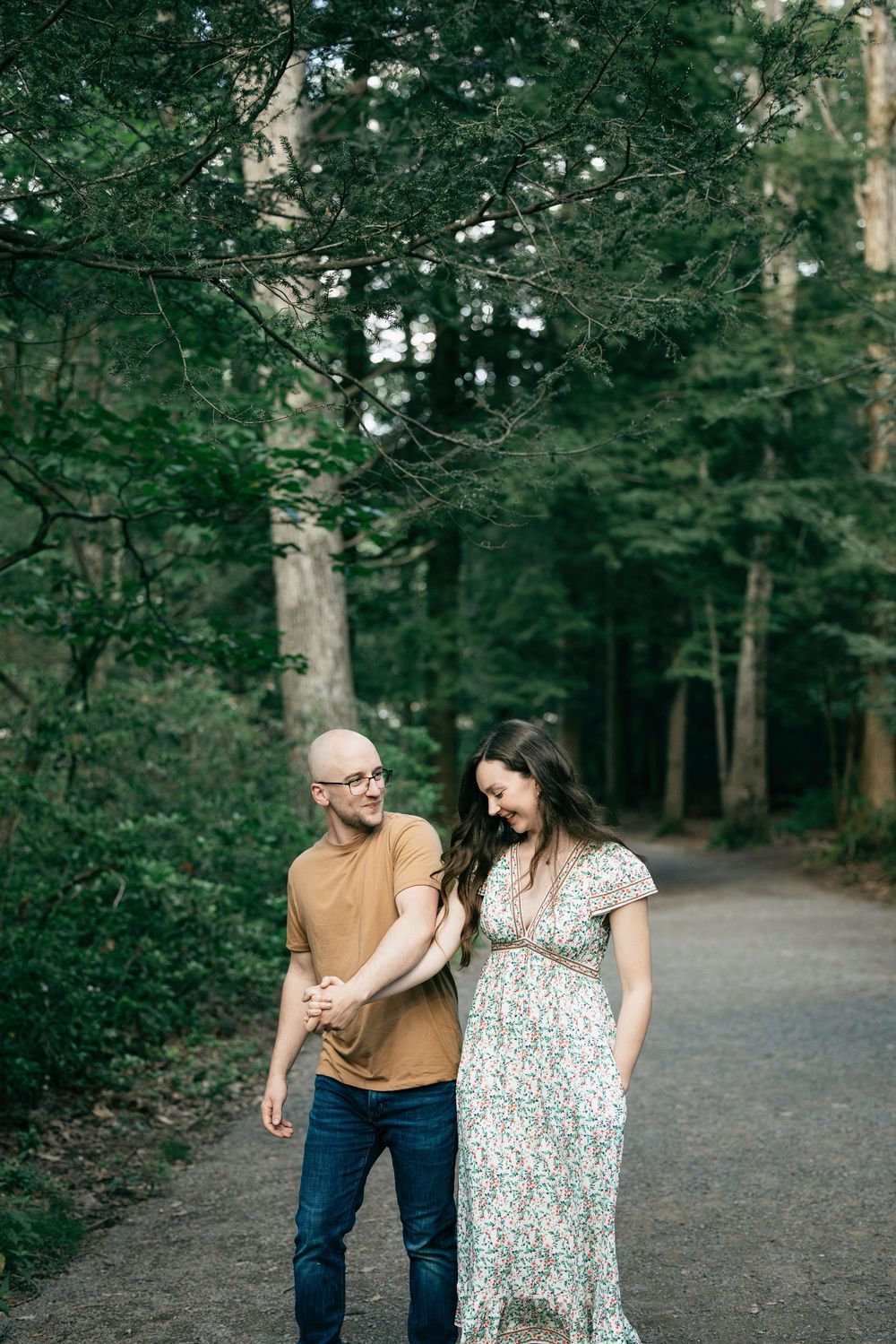 Pregnant couple holding hands on a forest path, man in tan shirt and jeans, woman in floral maxi dress.
