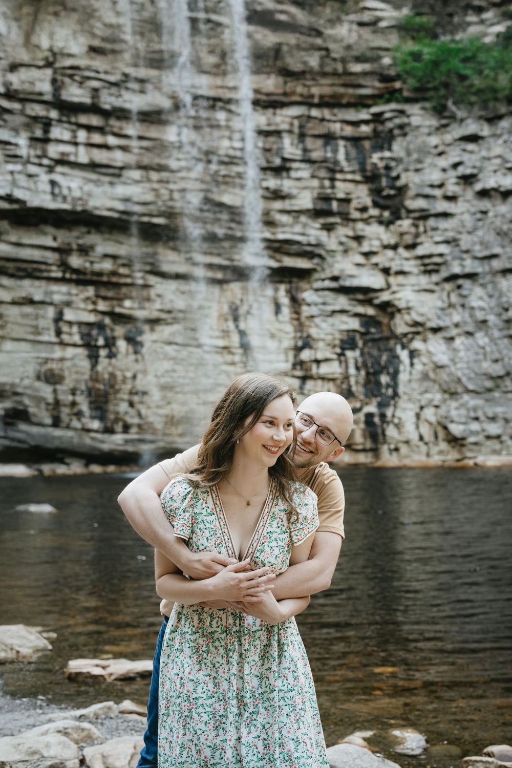 A couple embraces romantically in front of a stunning waterfall and rocky cliff, the woman in a floral dress smiling warmly.