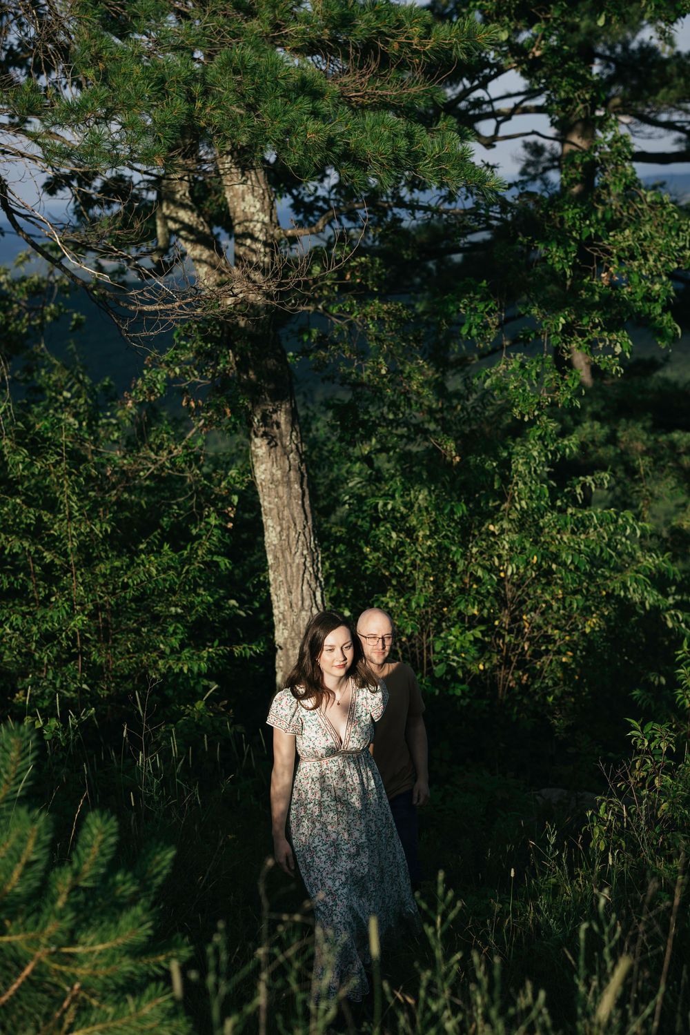 A couple stands together among lush green trees and foliage in a serene outdoor forest setting.