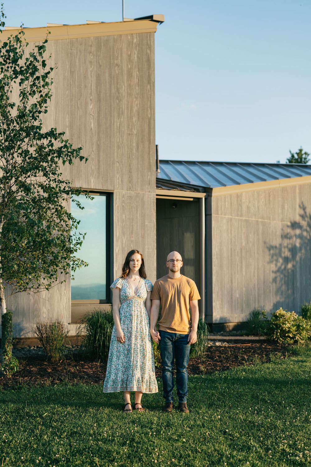 A couple stands on green grass in front of a modern wood and metal home during golden hour.