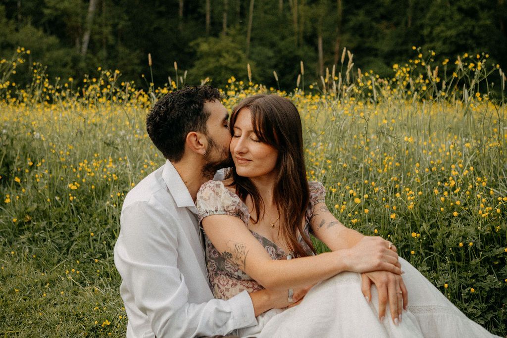 Couple sitting in a wildflower meadow, man kissing woman on cheek as she smiles softly.