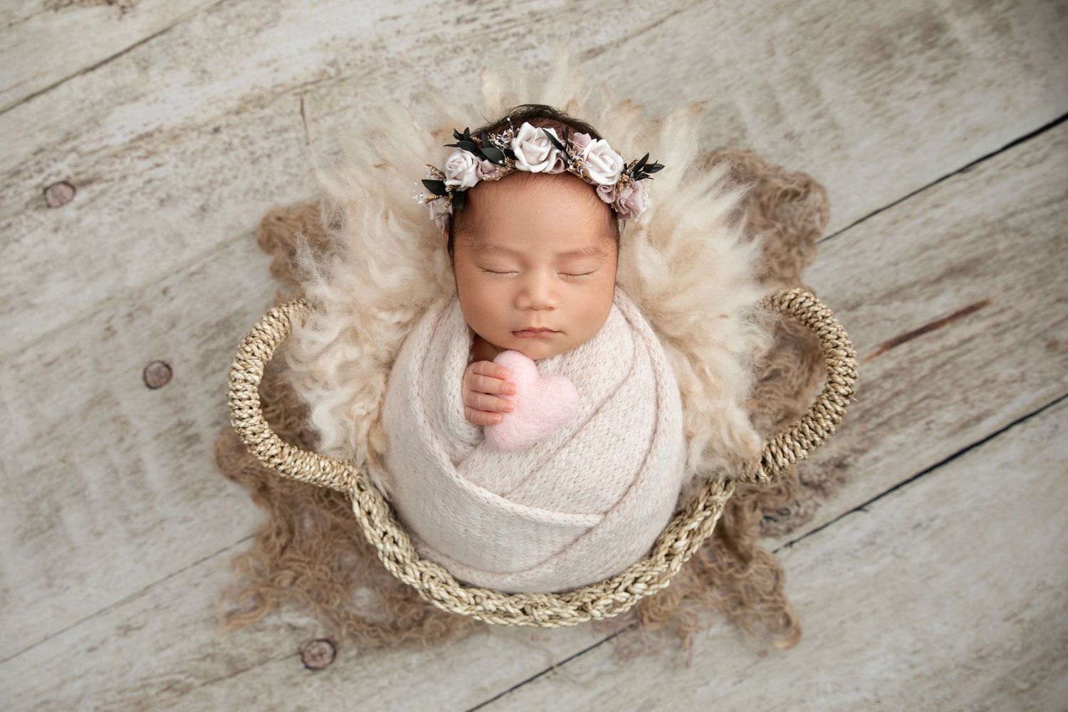 Newborn baby wrapped in cream swaddle with floral crown sleeping in wicker basket on rustic wood background.