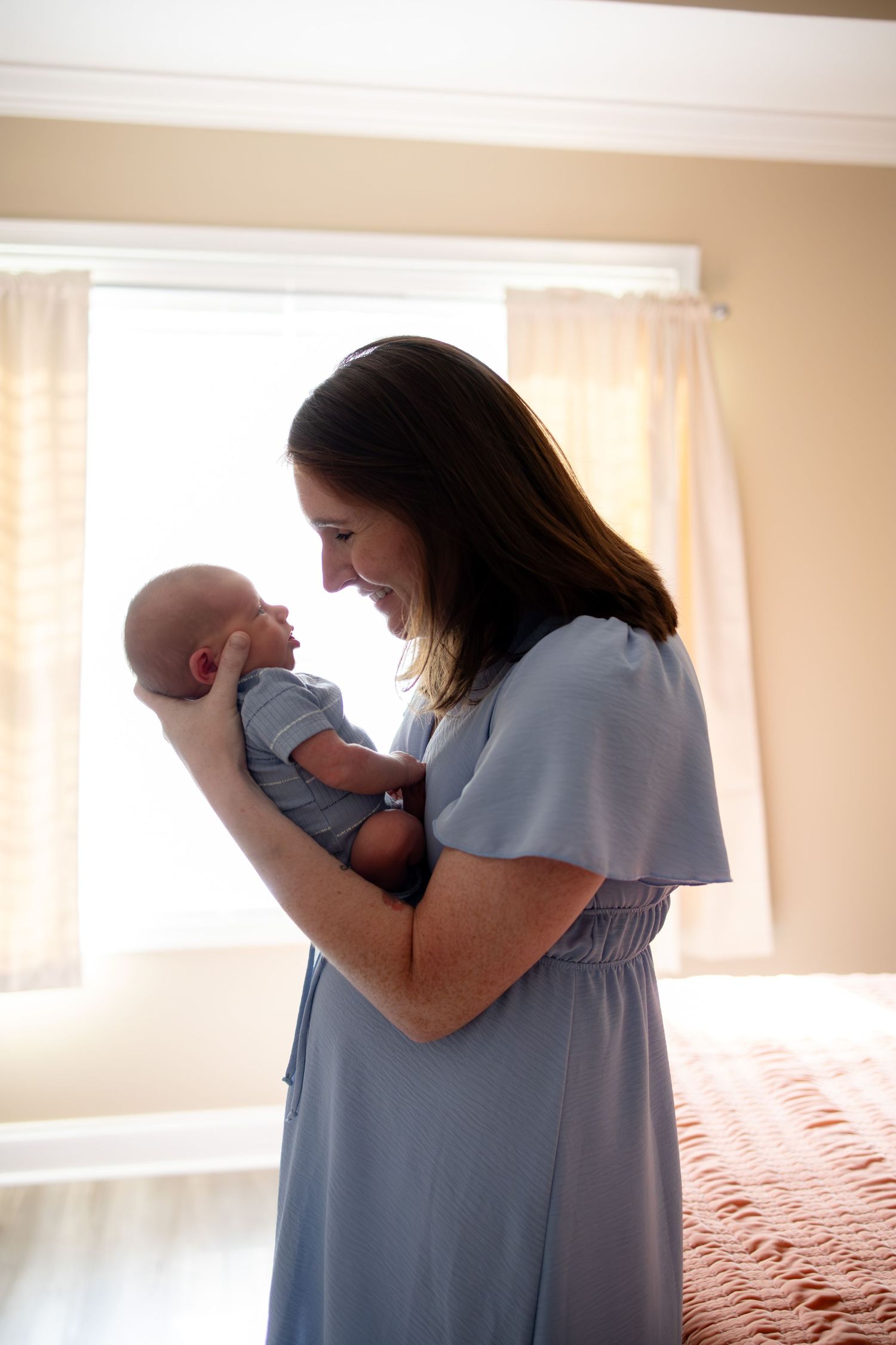 A smiling mother in a blue dress tenderly holds her newborn baby in a sunlit bedroom.