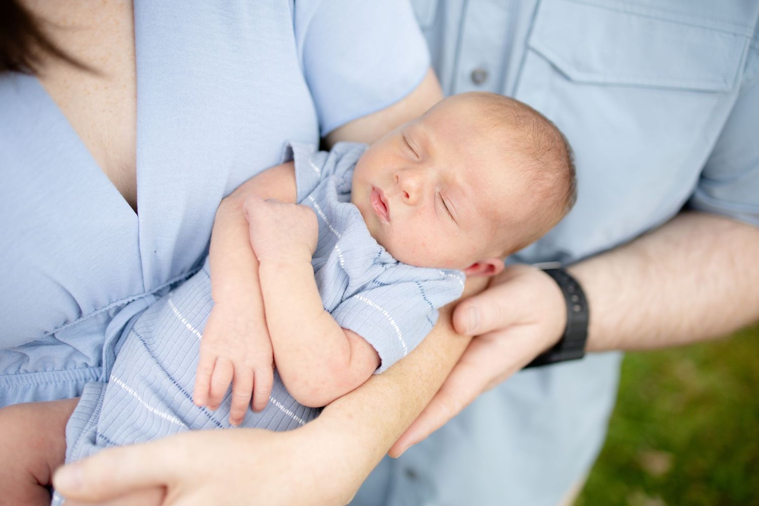 Parents in blue outfits gently cradle their sleeping newborn baby outdoors on green grass.