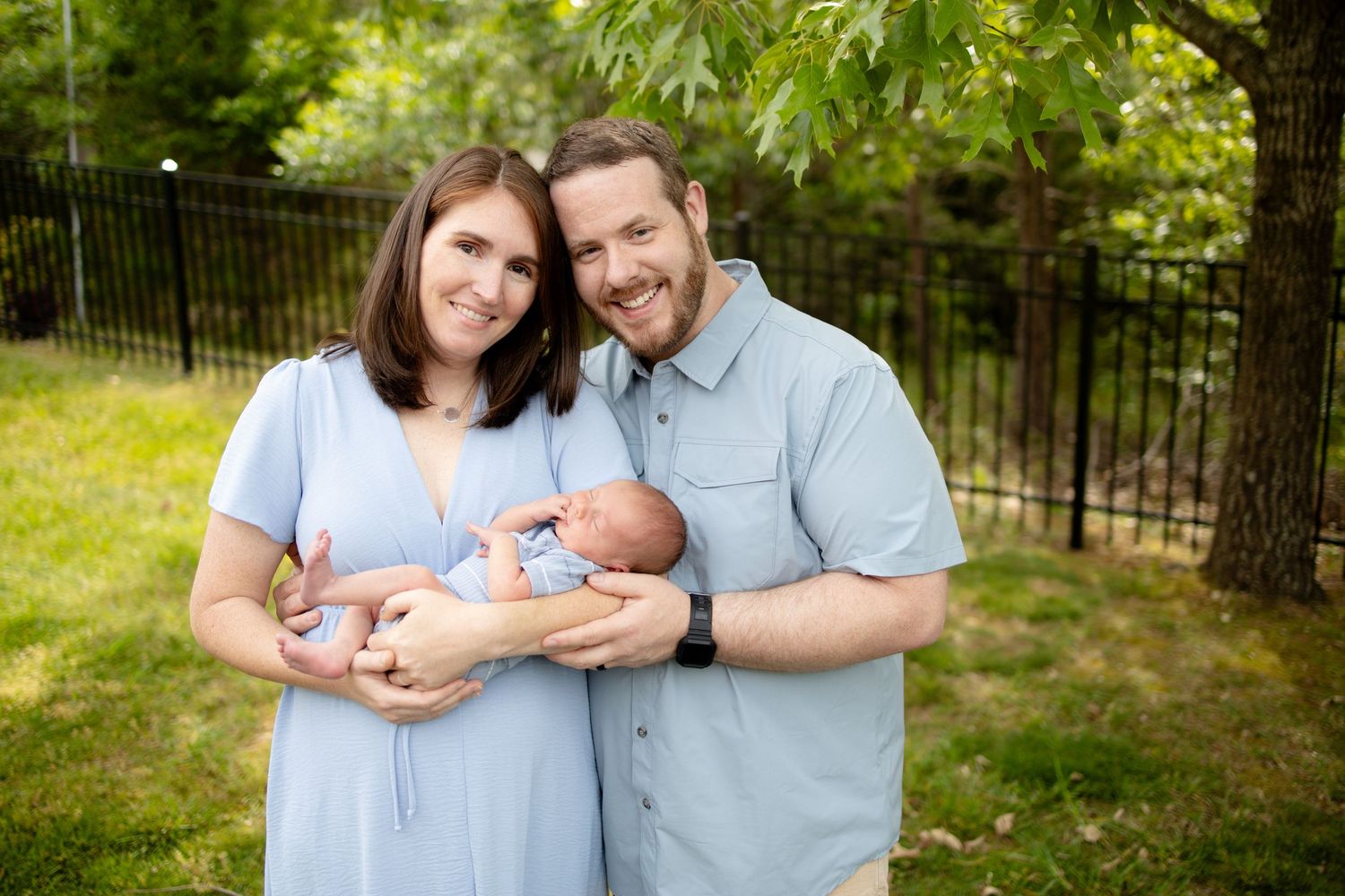 Smiling parents holding their newborn baby together in a lush green backyard garden setting.