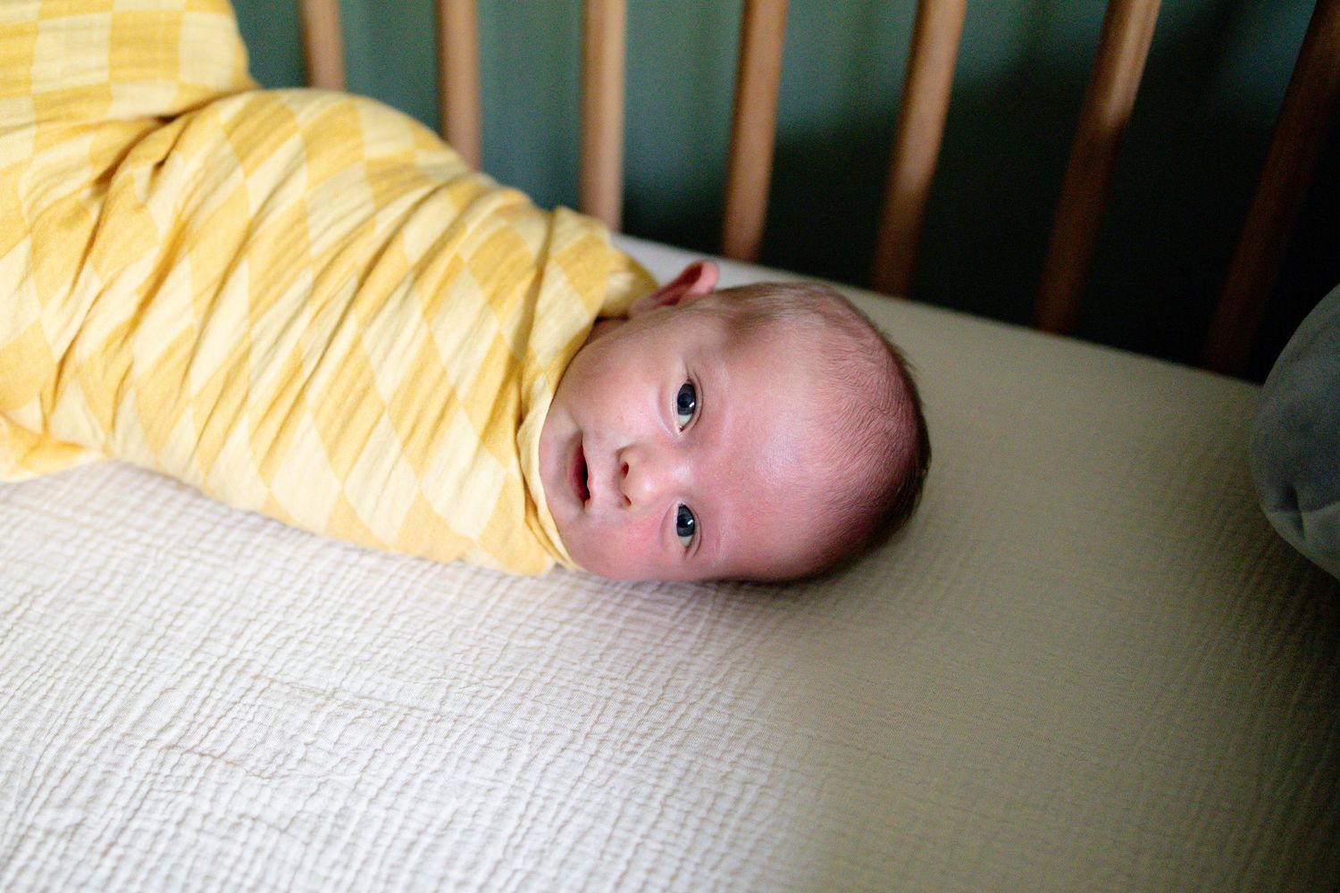 Newborn baby swaddled in yellow blanket lies in a wooden crib with wide eyes.