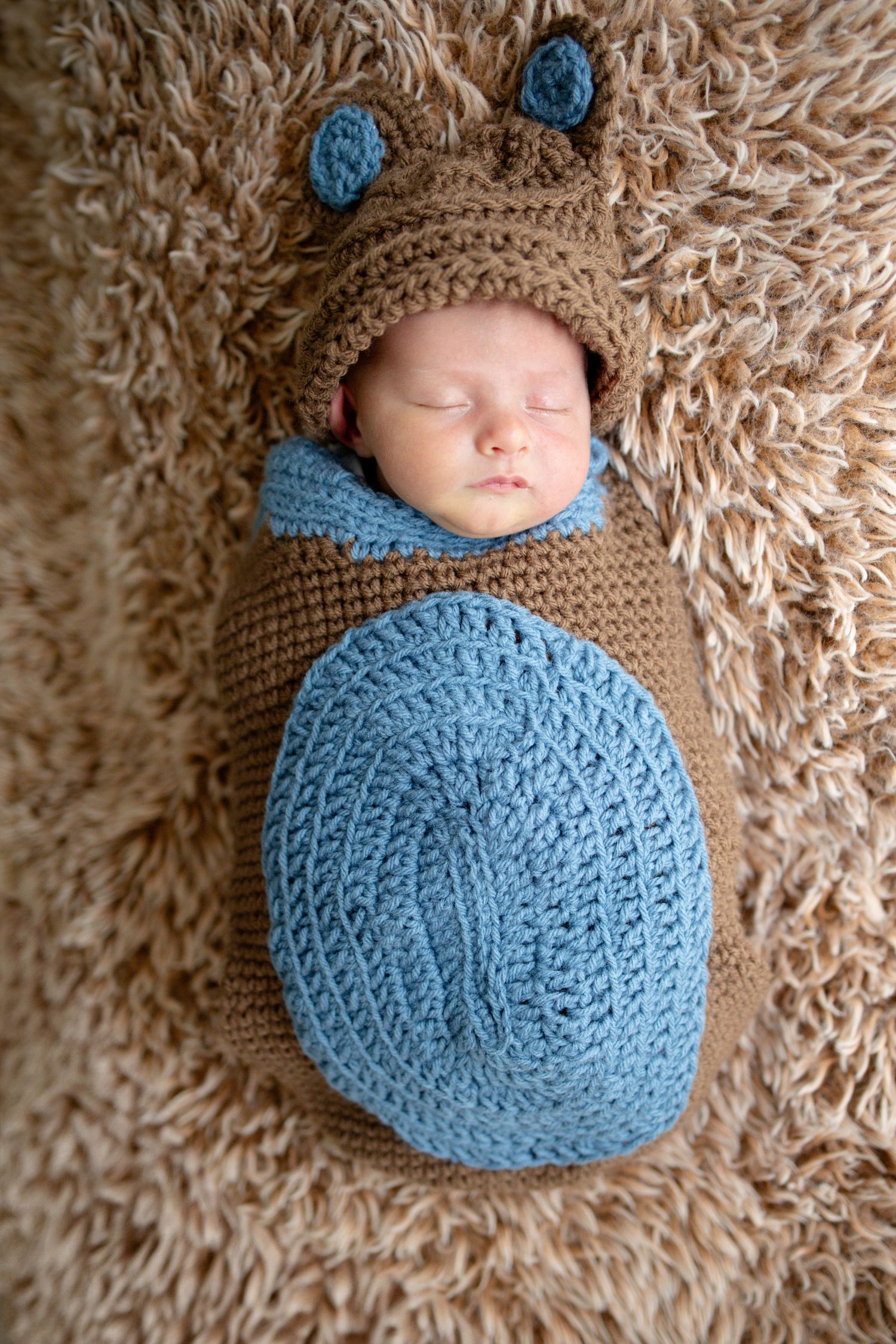 Sleeping newborn baby swaddled in a crocheted brown and blue bear cocoon on fluffy rug.