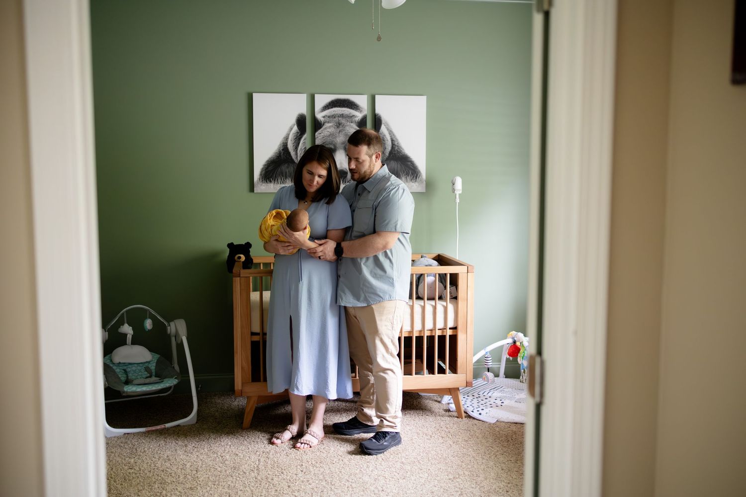 New parents cradle newborn in a cozy green nursery, viewed through a doorway.
