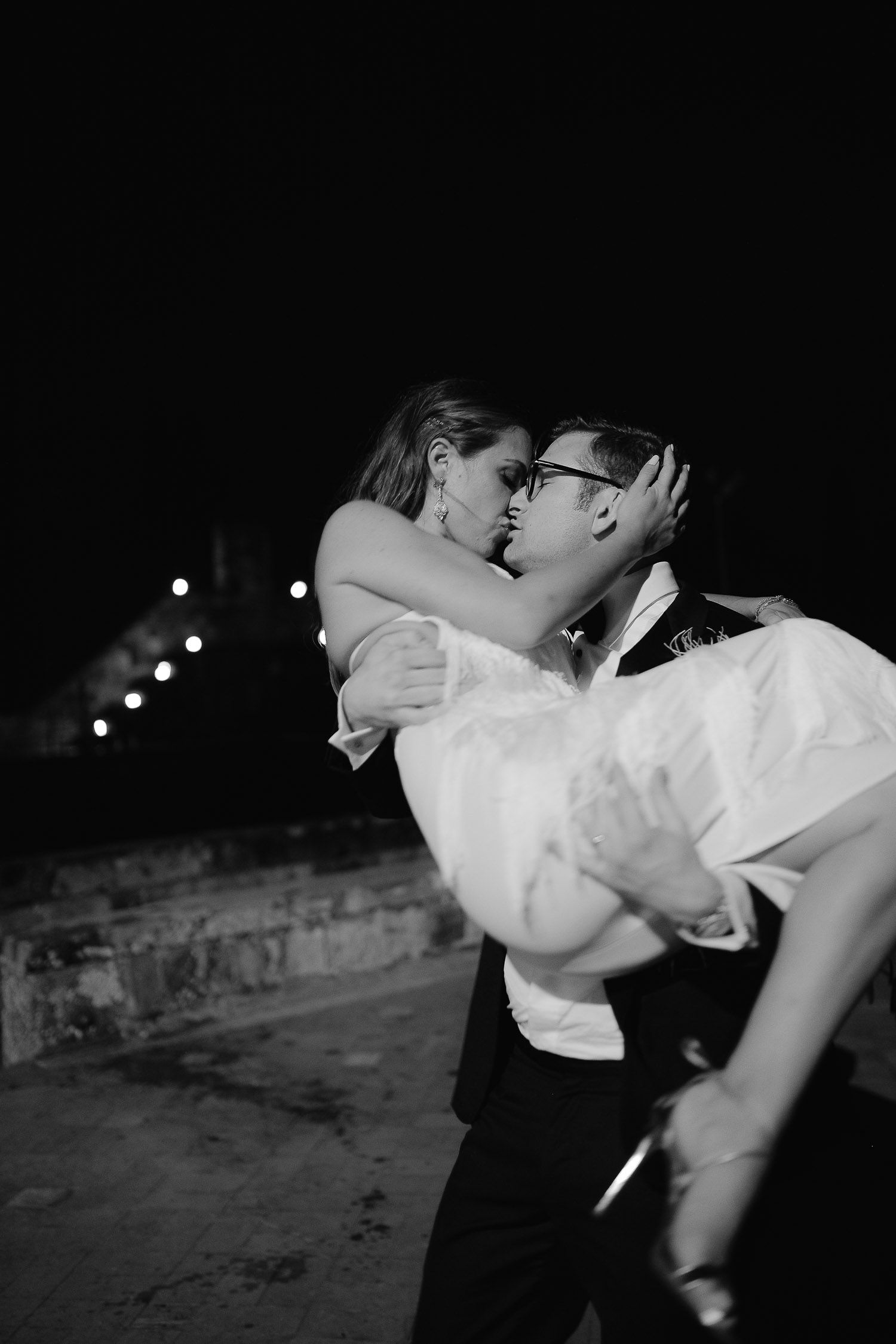 Black and white photo of a couple sharing a romantic kiss outdoors at night, man lifting woman in formal attire.