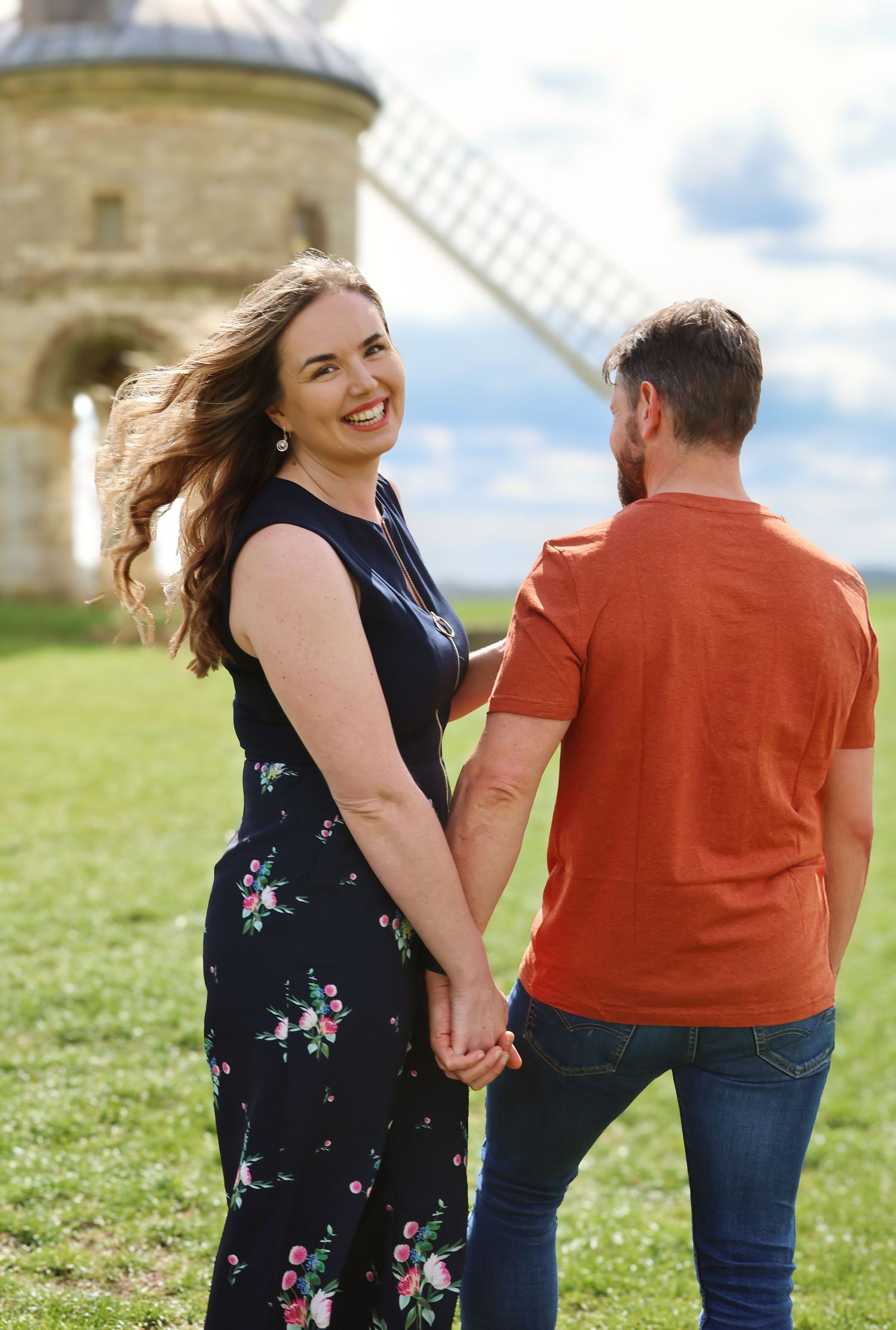 Couple holding hands near a historic windmill on a sunny day, woman in floral dress smiling over shoulder.