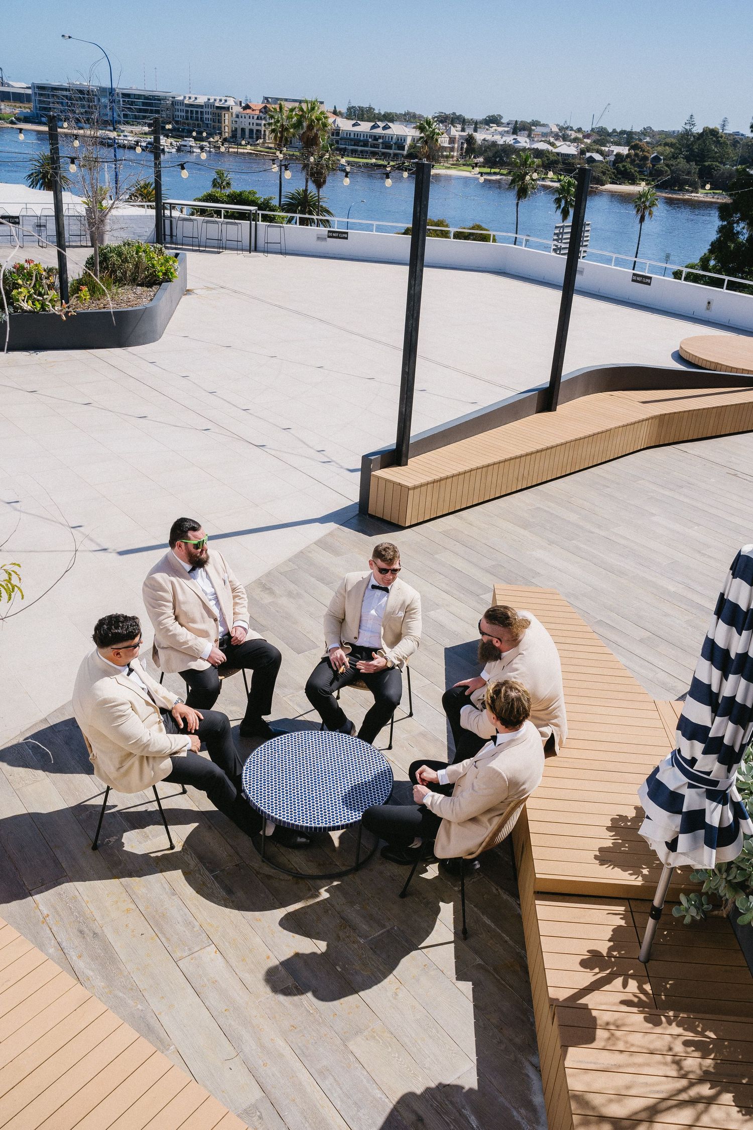 Groomsmen in cream suits gather around a table on a waterfront rooftop deck on a sunny wedding day.