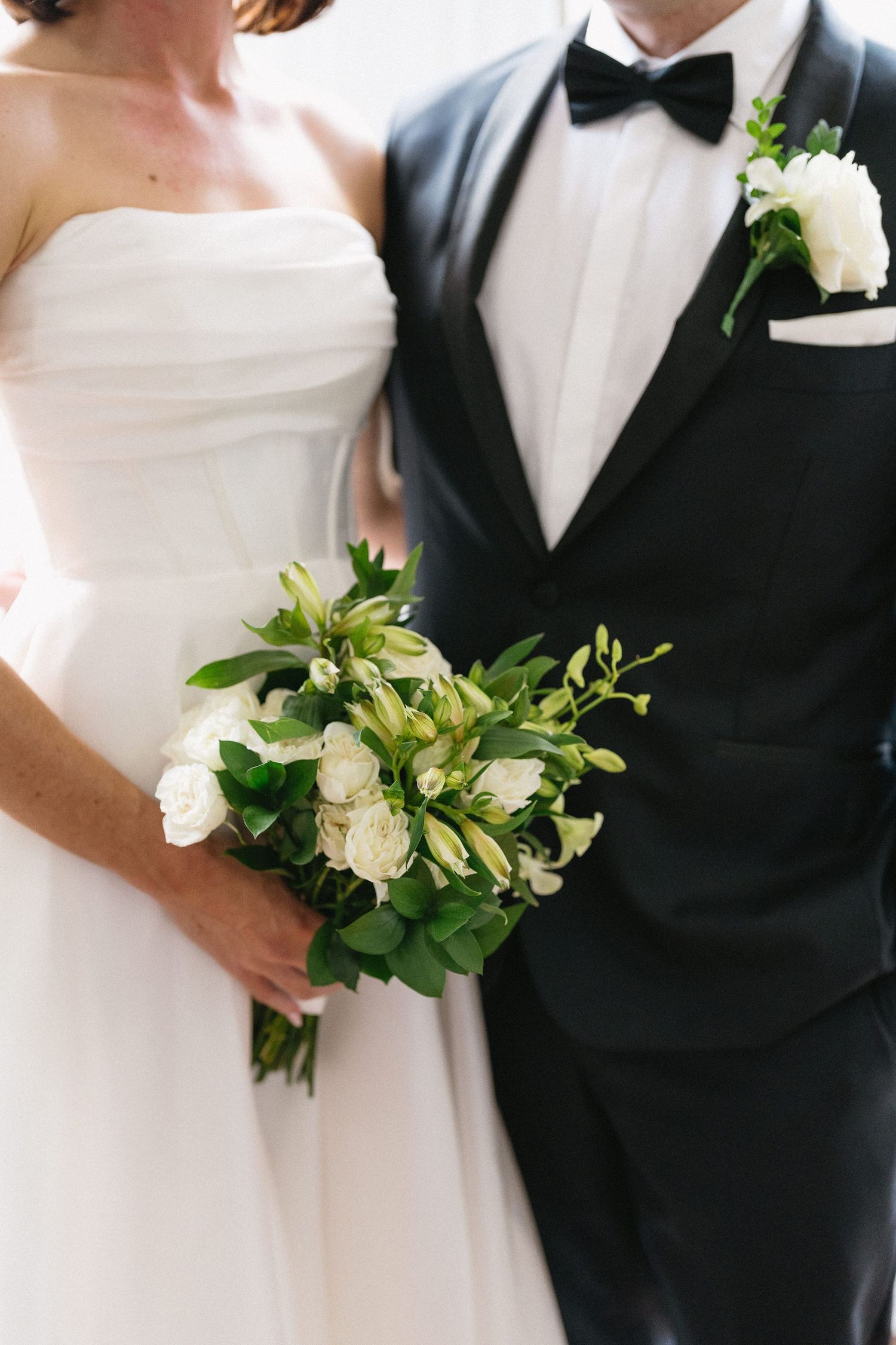Bride in strapless white gown holds green and white bouquet beside groom in black tuxedo with bow tie and boutonniere.
