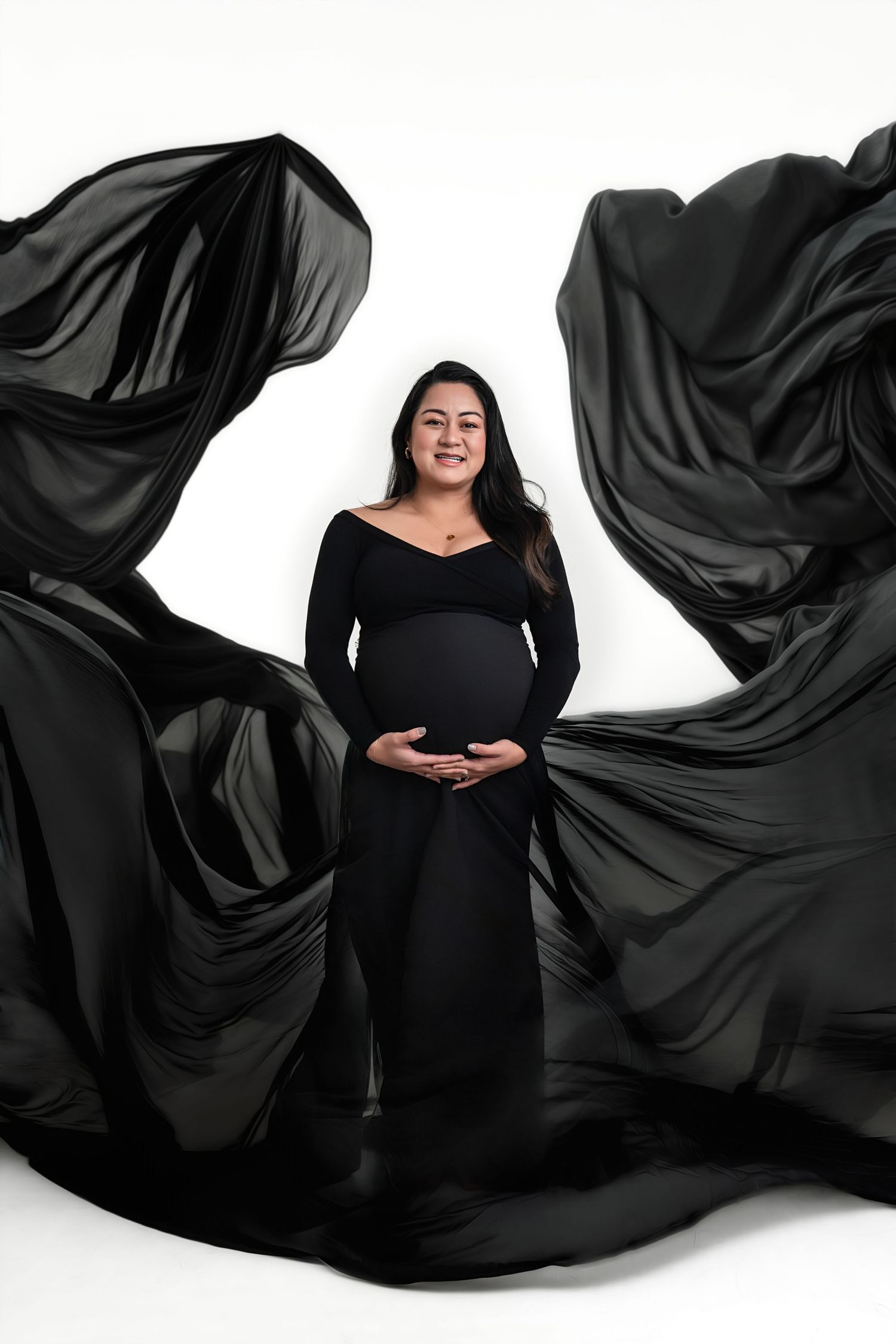 Pregnant woman in black maternity gown with flowing fabric wings poses against white studio background.