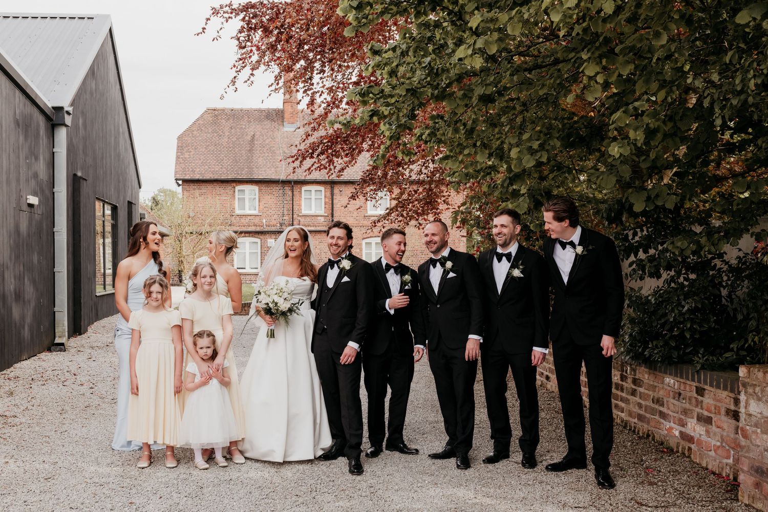 Wedding party poses outdoors, bride in white gown, groom and groomsmen in black tuxedos, bridesmaids in blush dresses.