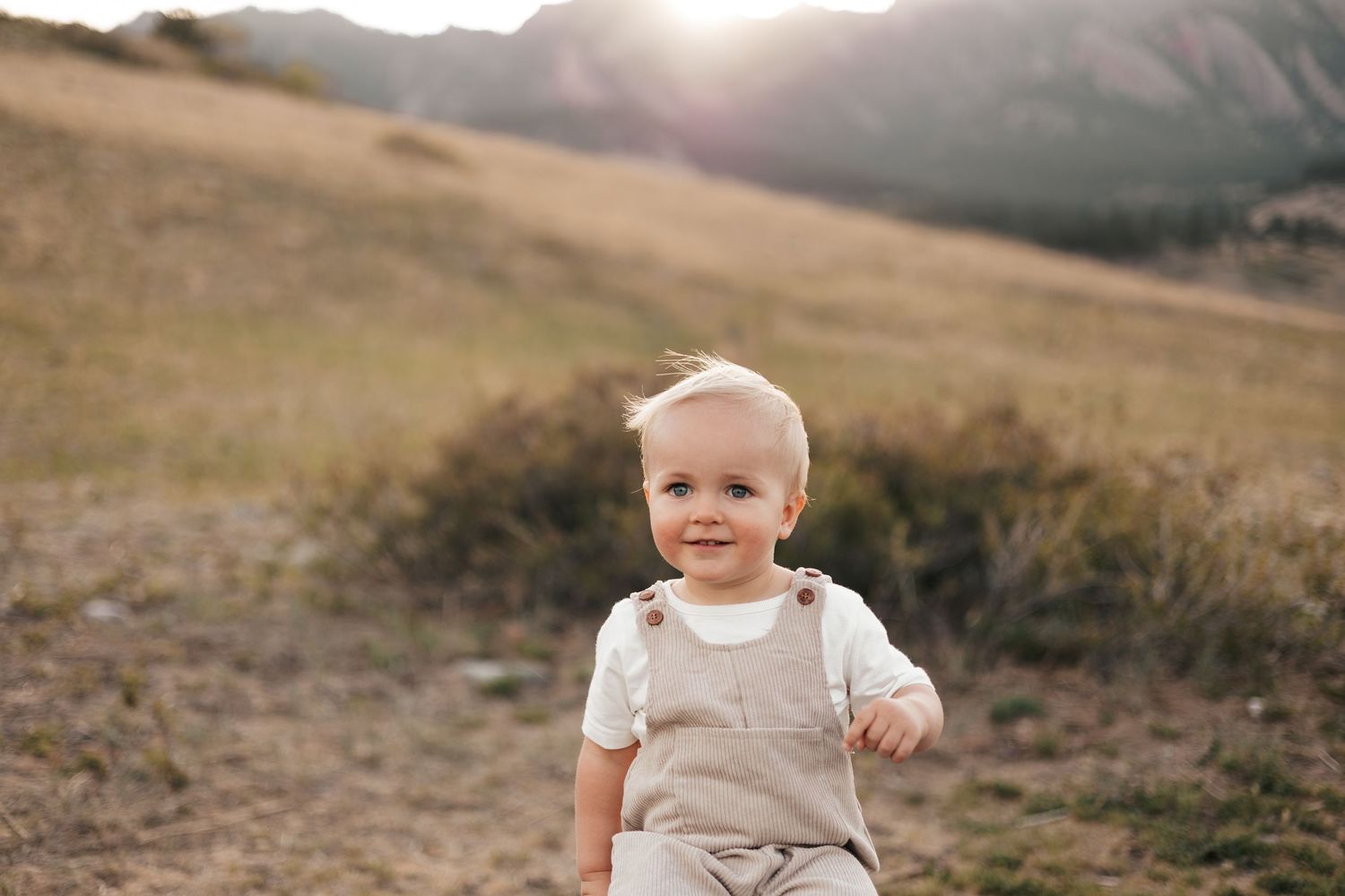 Smiling pregnant woman in floral dress with wind-blown hair poses outdoors with mountain landscape at golden hour.