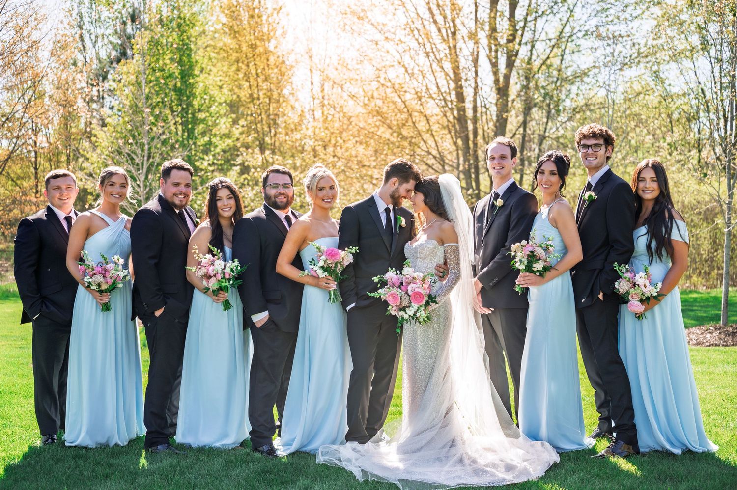 Bride and groom share romantic embrace in front of large wooden-framed windows with lush green trees in background.