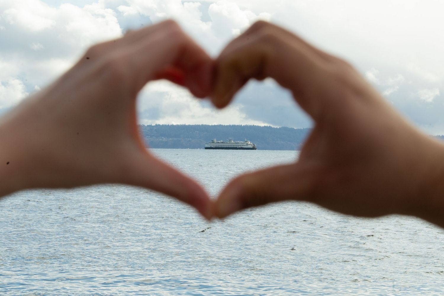 Hands forming a heart shape framing a large ship on calm blue ocean waters under a cloudy sky. Captured by Savvy Photography.