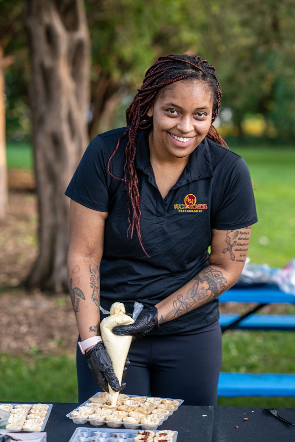 Smiling woman with tattoos and black gloves pipes frosting onto cupcakes outdoors at a catering event.