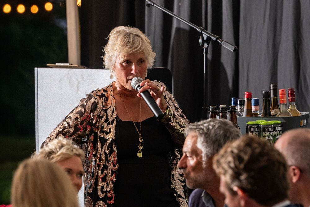 Woman speaking into microphone at outdoor evening event with bottles displayed in background.