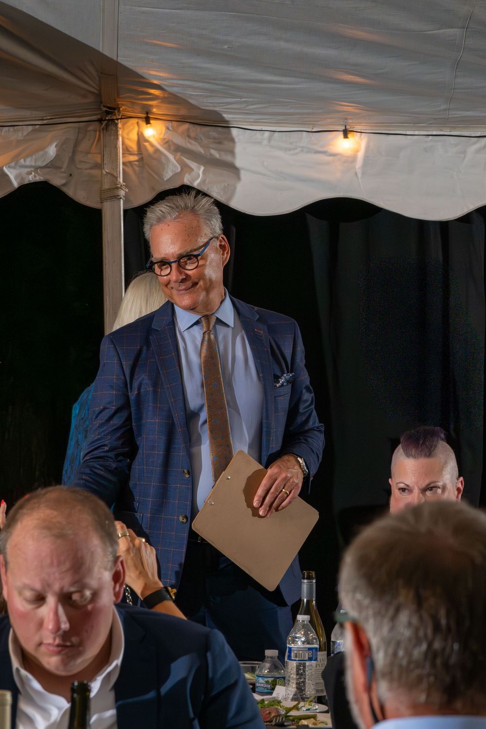 Smiling man in blue suit holding clipboard stands at elegant outdoor tent dinner event with seated guests.