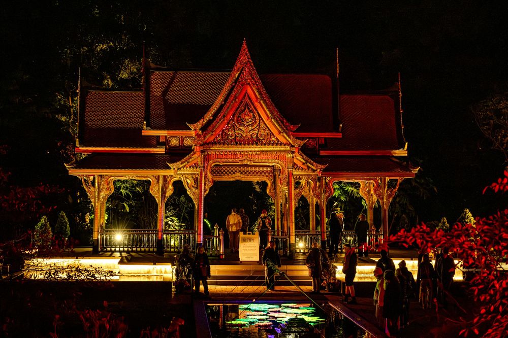 Illuminated Thai temple at night with golden architecture, red decorations, and colorful lights reflecting on water.