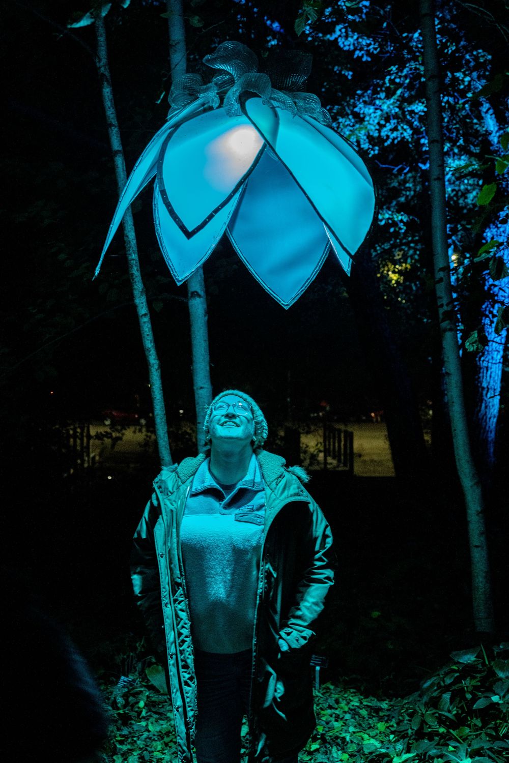 Person gazing up at a glowing blue lotus flower light installation among trees at night.