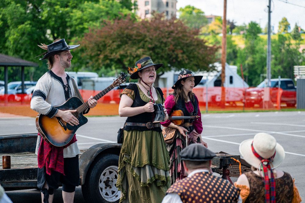 Three performers in Renaissance faire costumes play guitar and mandolin outdoors at a festival.