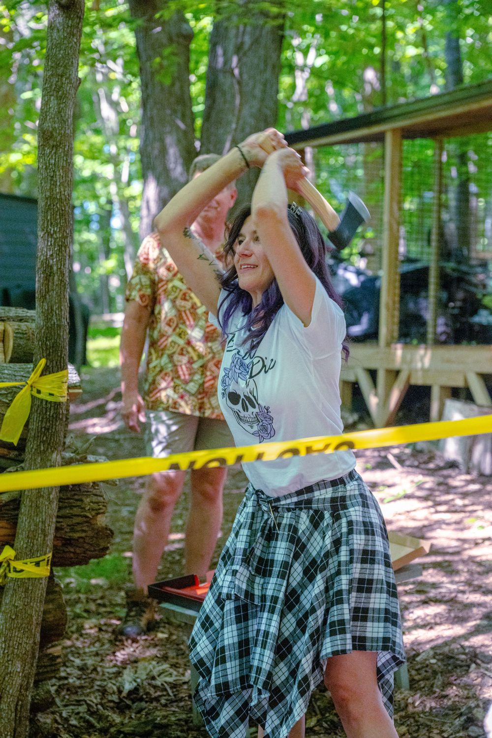 Woman with purple hair in plaid skirt throwing an axe outdoors at a woodland axe throwing range.