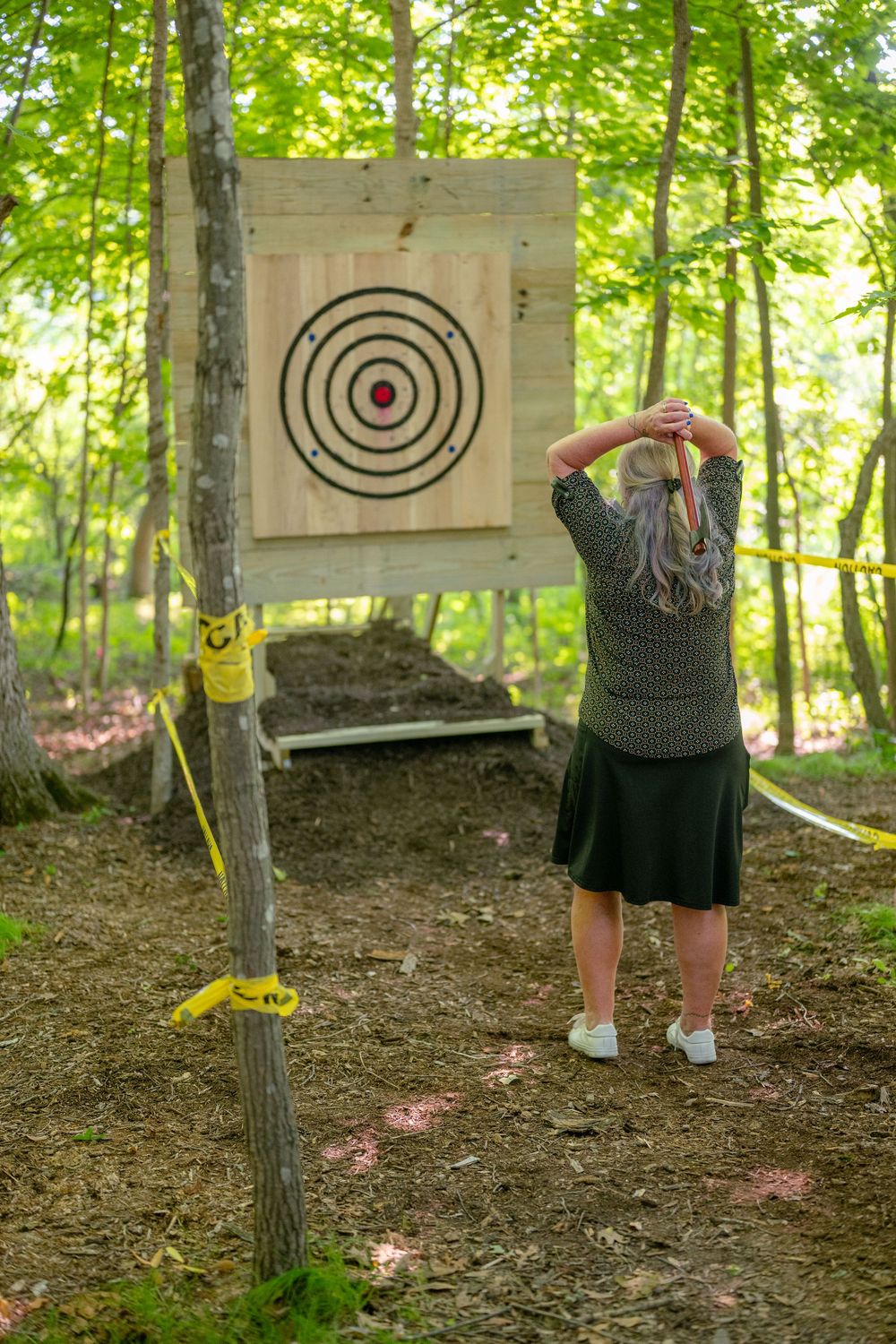 Woman with colorful hair preparing to throw an axe at a wooden target in an outdoor forest setting.