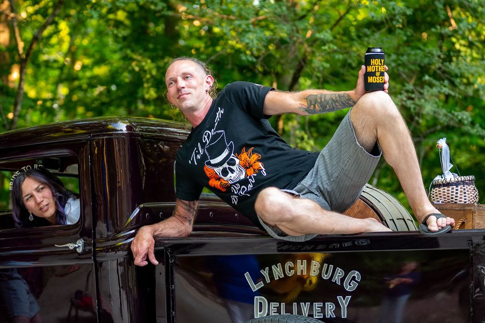 Man posing on a Lynchburg Delivery vintage truck holding a drink, with a woman leaning out the window outdoors.