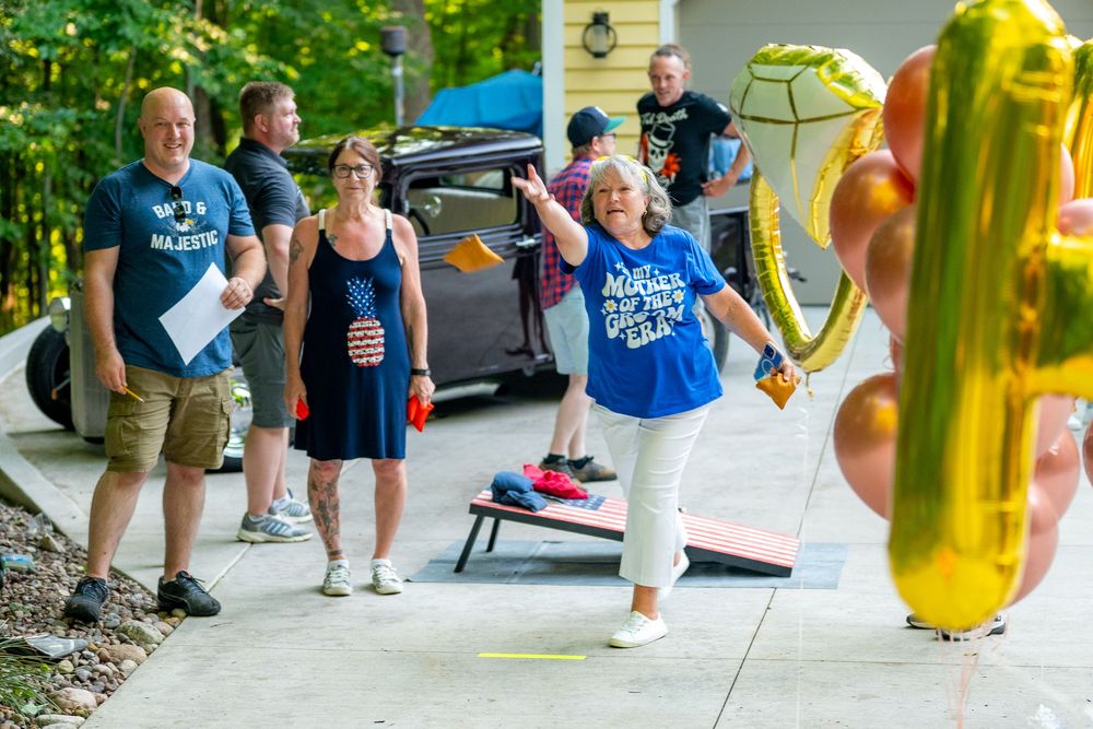 Woman in blue shirt waves holding gold balloons at outdoor party with guests on driveway near cornhole game.