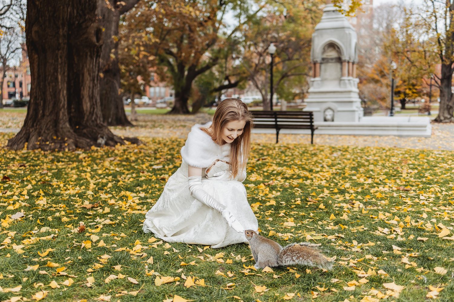 Bride in white gown feeding a squirrel in an autumn park with yellow fallen leaves and a monument in background.