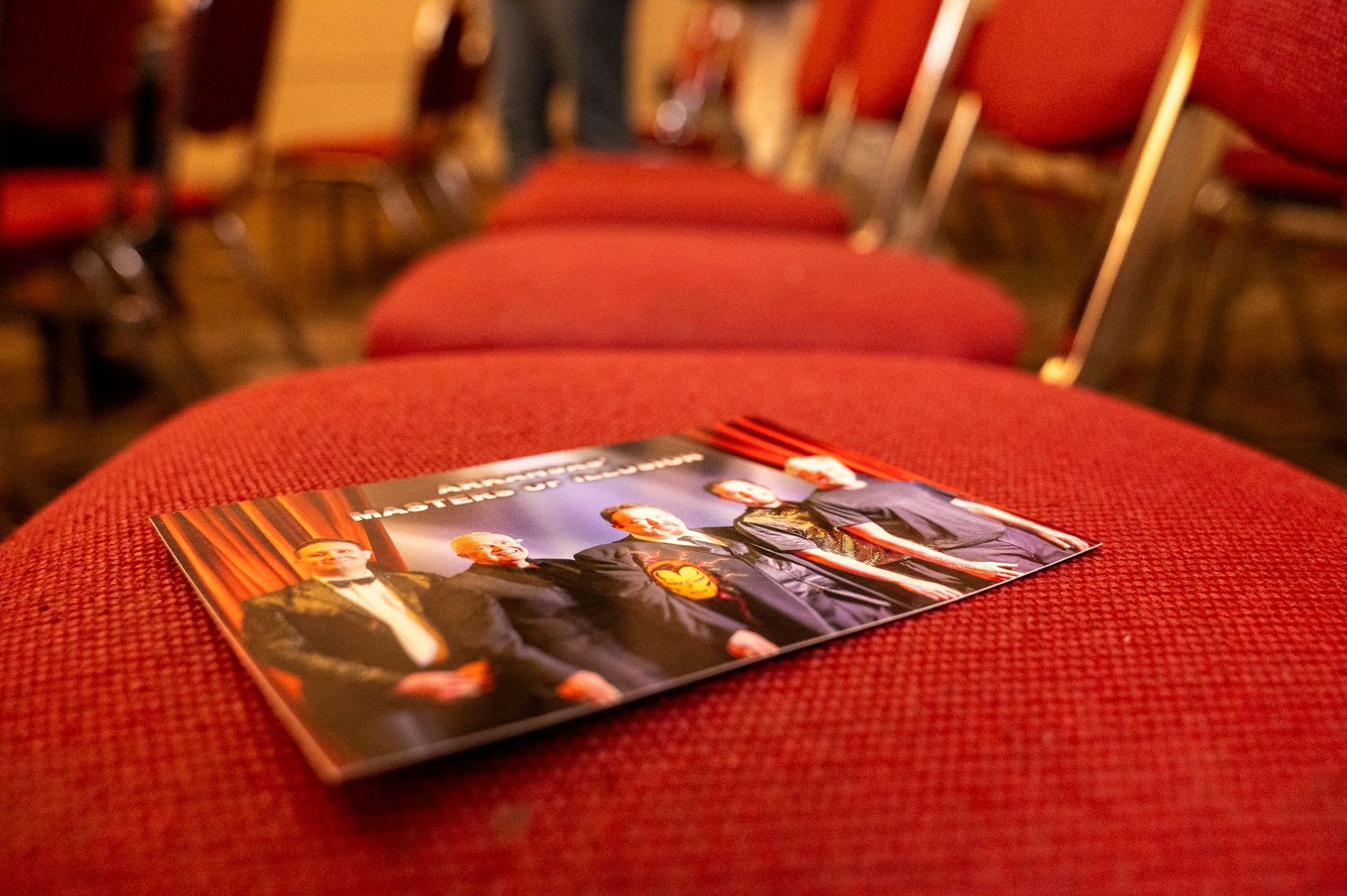 A program booklet resting on red cushioned chairs in an auditorium or conference venue setting.