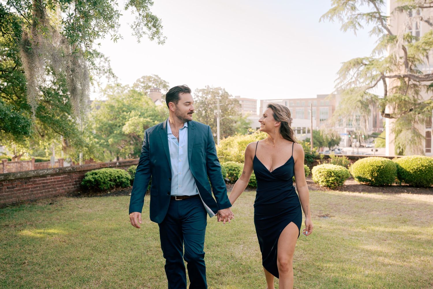 A couple in elegant attire holds hands and smiles while walking through a lush green park on a sunny day.