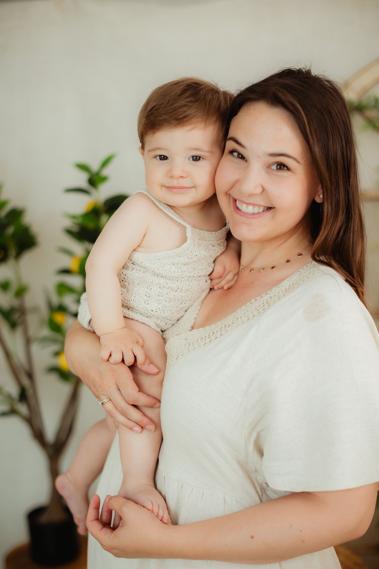 Smiling young mother in white dress holding her happy baby in a bright, plant-filled room.