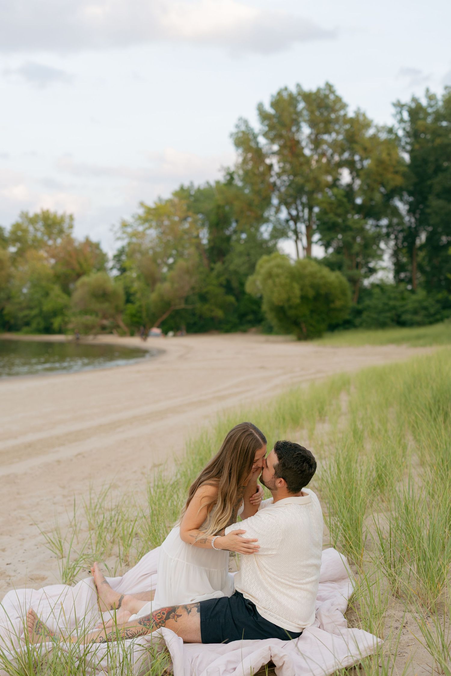 Couple sharing a romantic moment on a sandy beach, surrounded by green grass and trees at dusk.